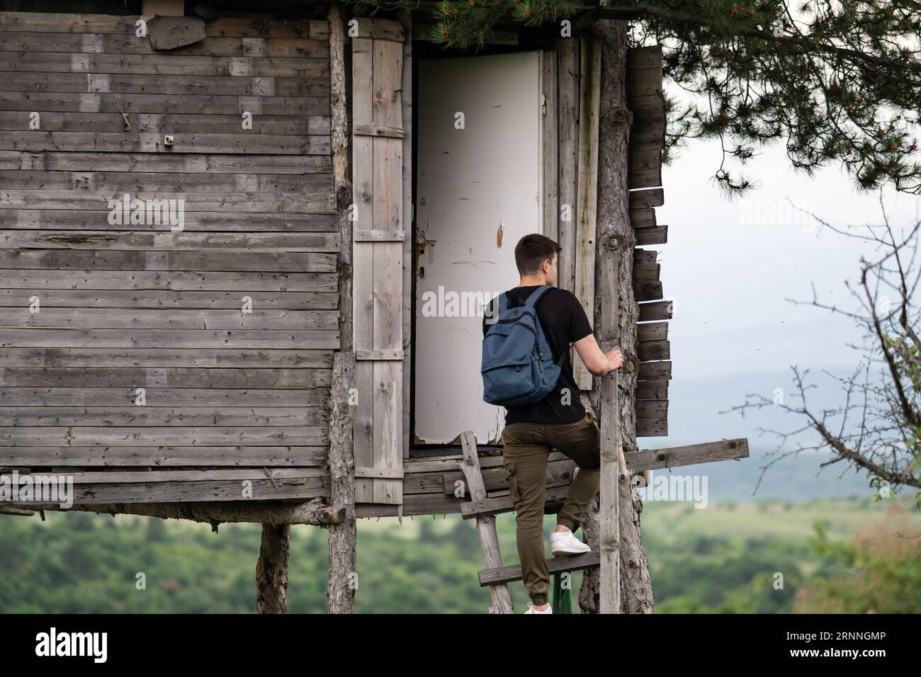 bellissimo giovane con zaino in piedi sulle scale di una casa di legno selvaggia e scattare foto con il suo cellulare. Escursionista di fronte alla casa sull'albero Foto Stock