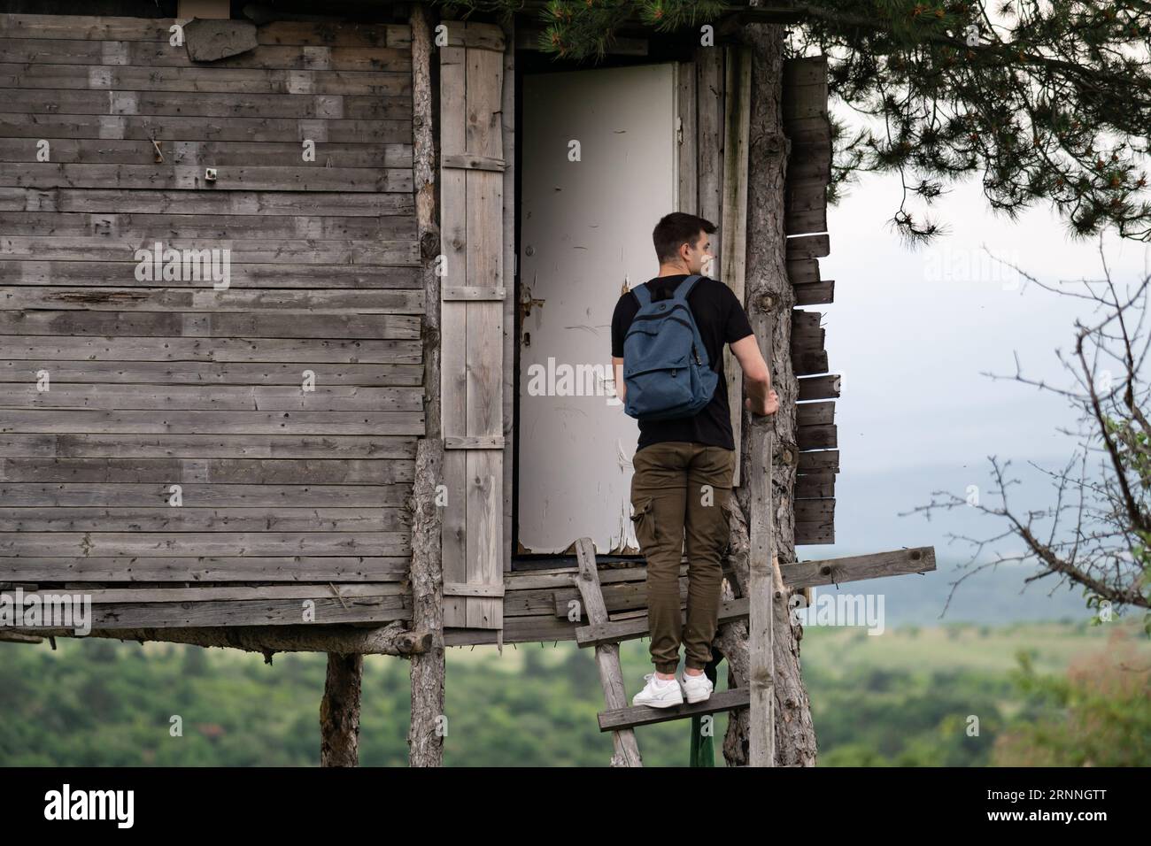 bellissimo giovane con zaino in piedi sulle scale di una casa di legno selvaggia e scattare foto con il suo cellulare. Escursionista di fronte alla casa sull'albero Foto Stock