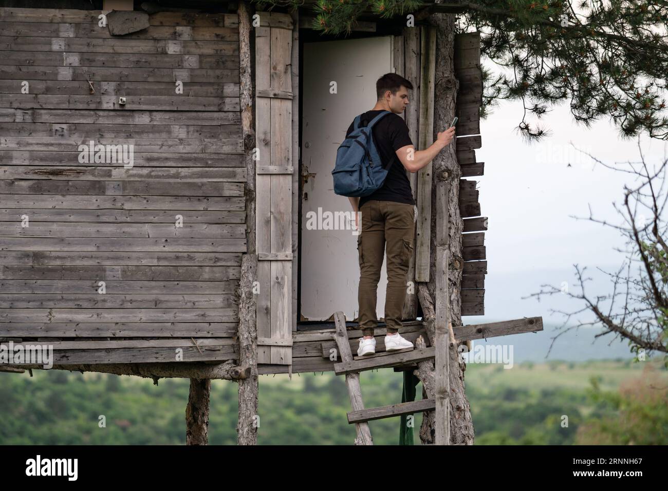 bellissimo giovane con zaino in piedi sulle scale di una casa di legno selvaggia e scattare foto con il suo cellulare. Escursionista di fronte alla casa sull'albero Foto Stock