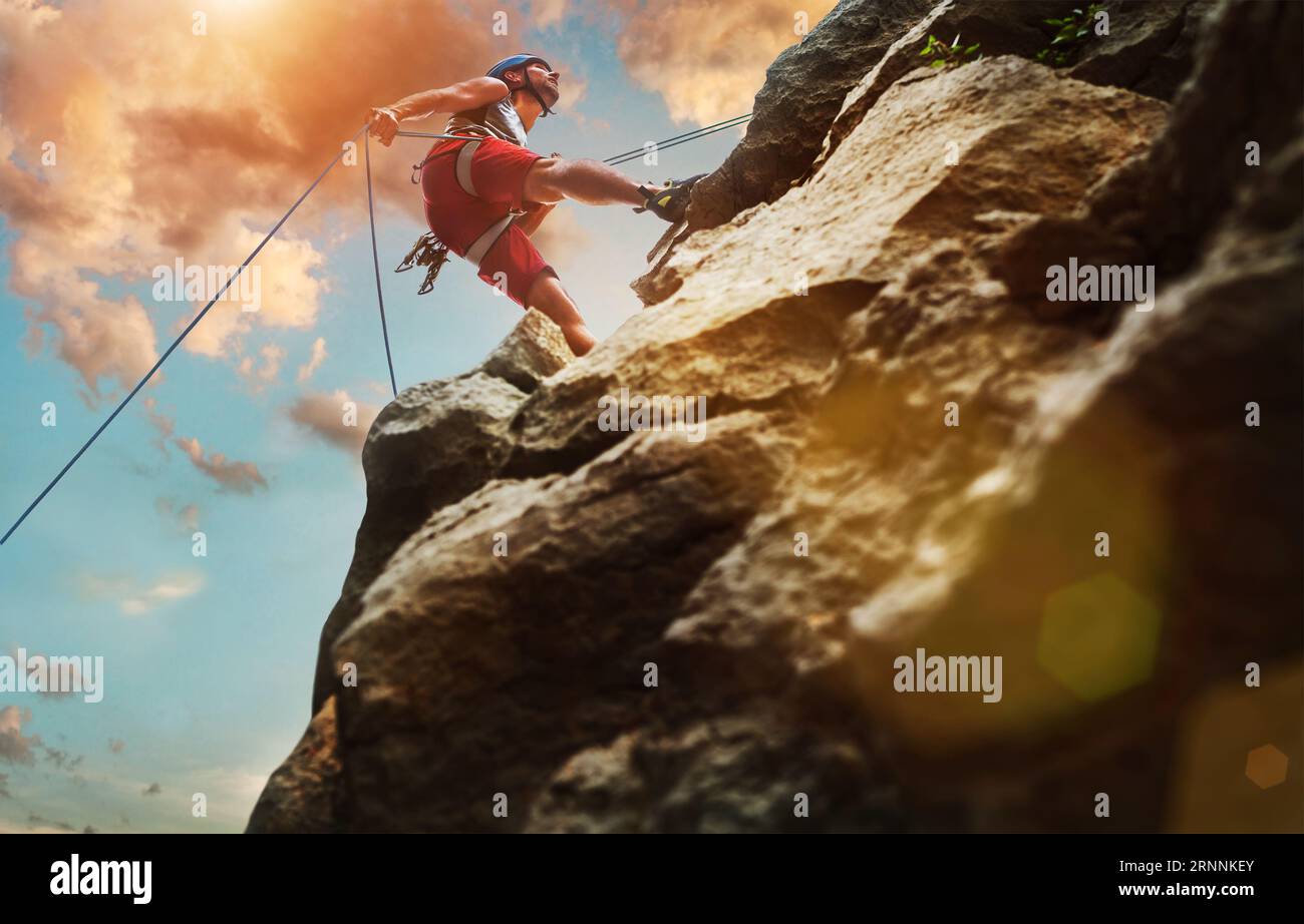Uomo climber muscoloso in casco protettivo che si abbella dalla parete rocciosa della scogliera utilizzando il dispositivo Belay corda e l'imbracatura di arrampicata su sfondo cielo tramonto sera Foto Stock