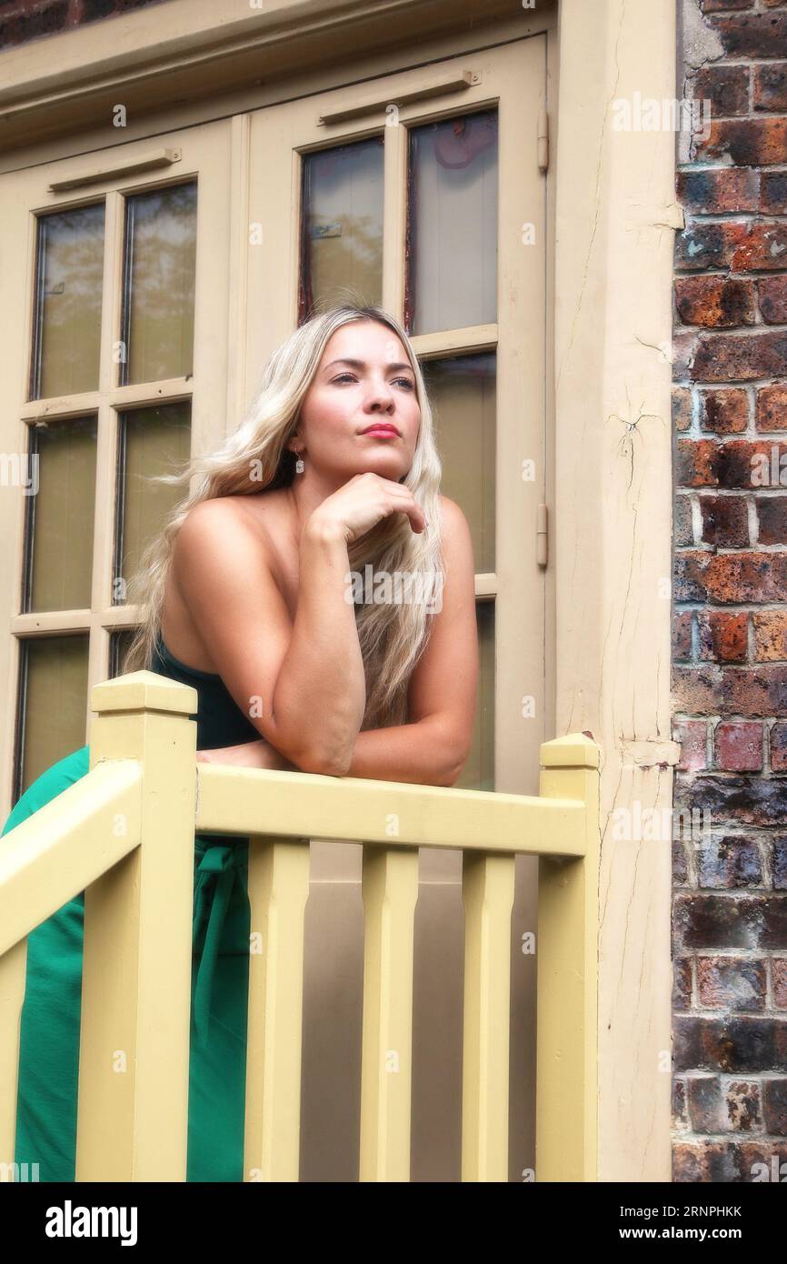 Donna bionda con capelli lunghi e vestito verde in posa davanti a una casa Foto Stock