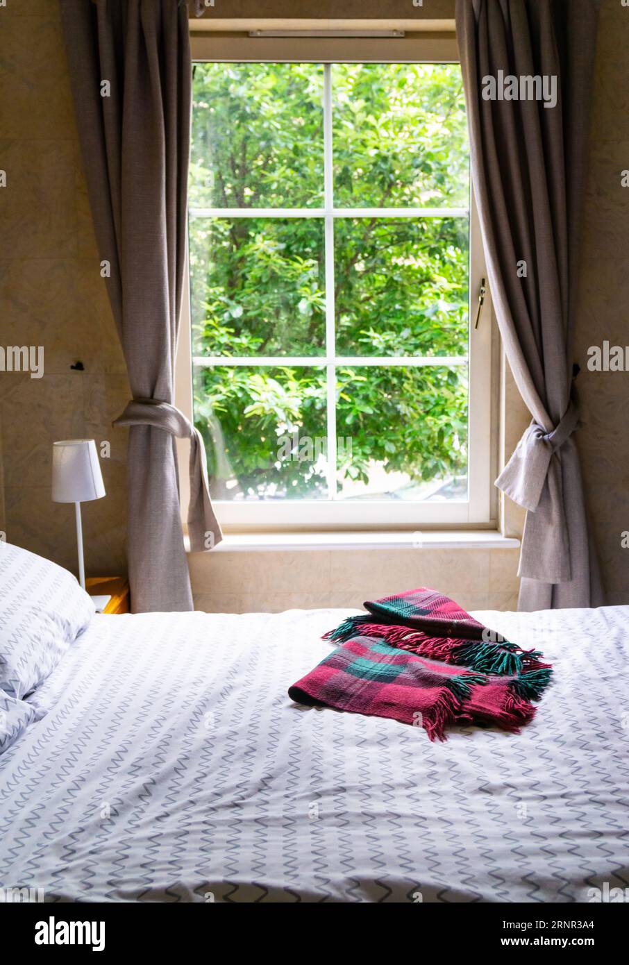 Vista parziale di una camera. Un letto con lenzuola bianche e una coperta scozzese in tartan sulla parte superiore, lampada da tavolo su un lato. Sullo sfondo una finestra con cornice bianca Foto Stock