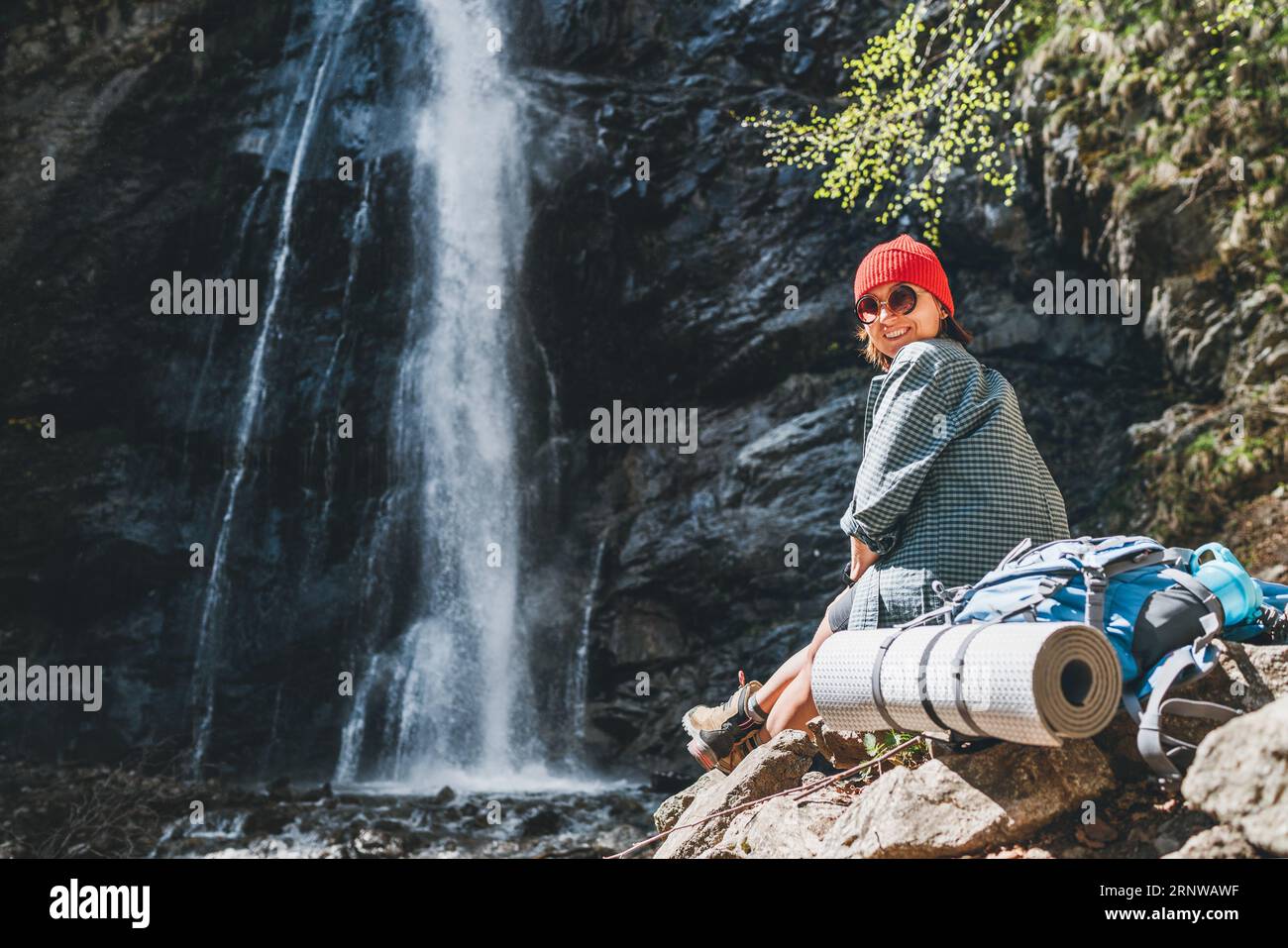 Donna sorridente con zaino in cappello rosso vestito con abiti da trekking attivi e stivali seduti vicino alla cascata del fiume di montagna e godersi gli spruzzi Natur Foto Stock