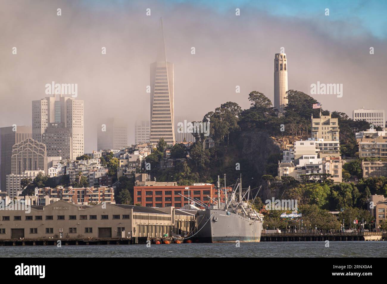 San Francisco, CA, USA - 12 luglio 2023: Paesaggio urbano sotto la nebbia marrone con la torre Coit nel Pioneer Park, Transamerica Pyramid con altri edifici. Foto Stock