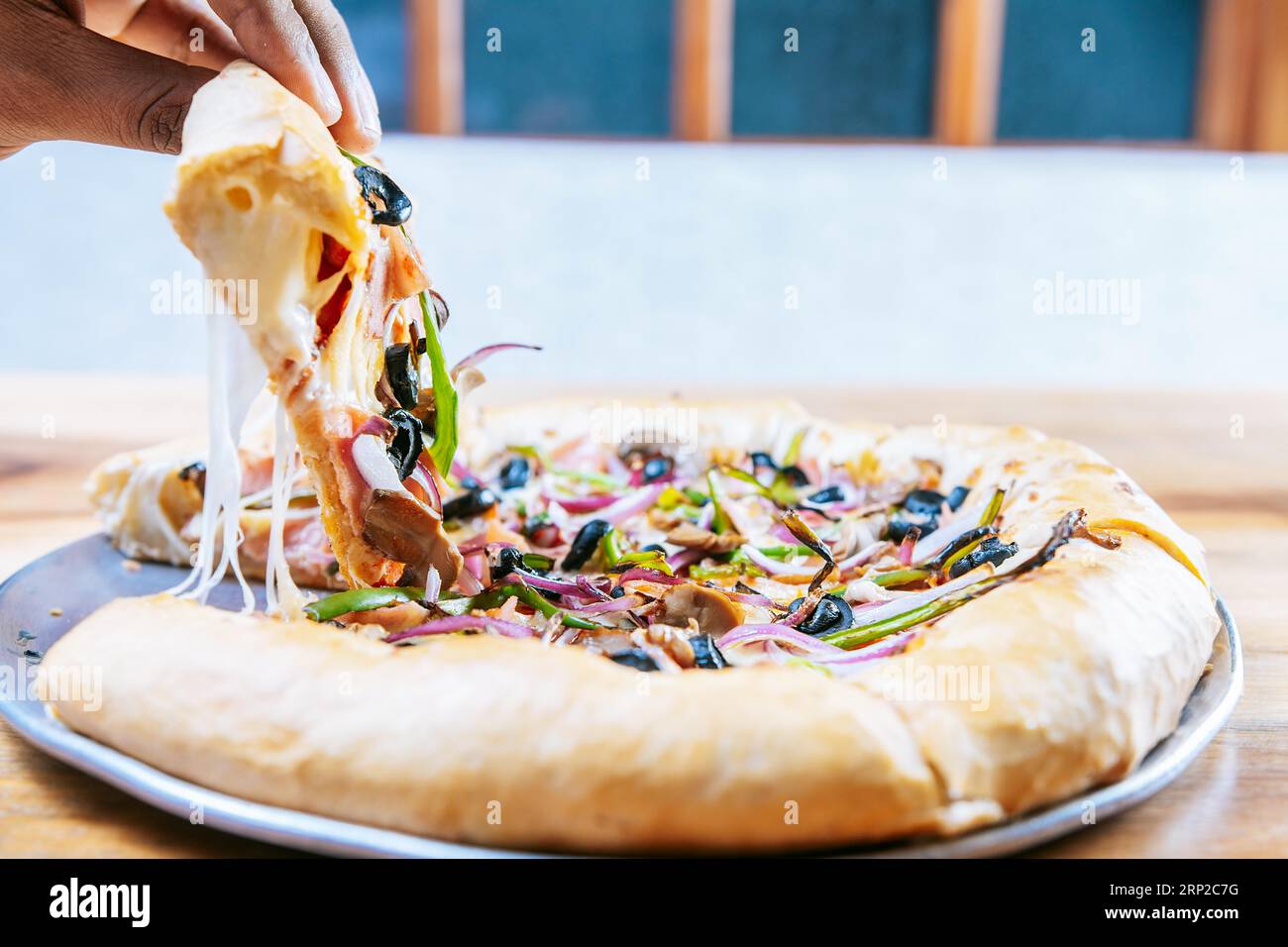 A mano si prende una fetta di pizza suprema su un tavolo di legno. Primo piano di una mano che prende una deliziosa fetta di pizza suprema con verdure Foto Stock