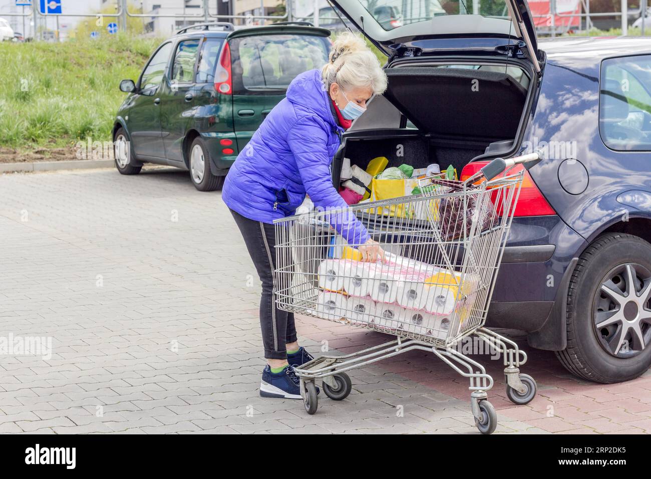 Una donna anziana dall'aspetto buono che indossa una maschera protettiva carica gli acquisti in un'auto nel parcheggio. Vita quotidiana durante la pandemia di coronavirus. Foto Stock
