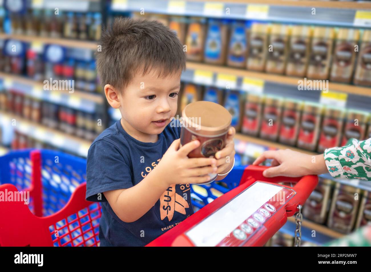 Adorabile bimbo multirazziale di due anni con una t-shirt blu seduto in un carrello della spesa con generi alimentari, che tiene in mano una tazza di plastica con le mani piccole, in g Foto Stock