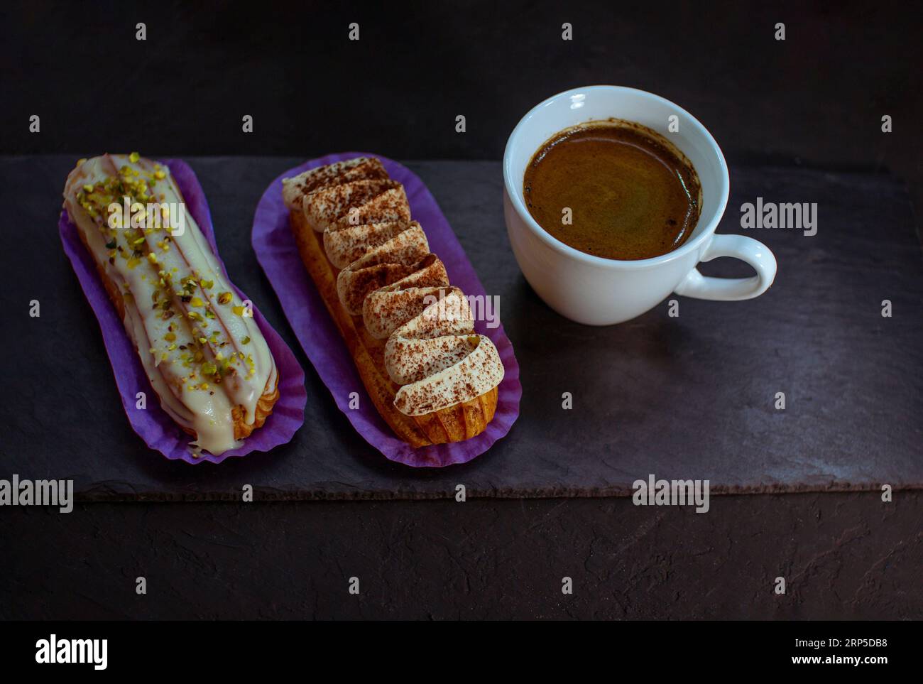 Tazza di caffè e deliziose torte eclair - tiramisù e pistacchio aromatizzati con panna, glassa e noci tritate, su fondo nero. Foto Stock