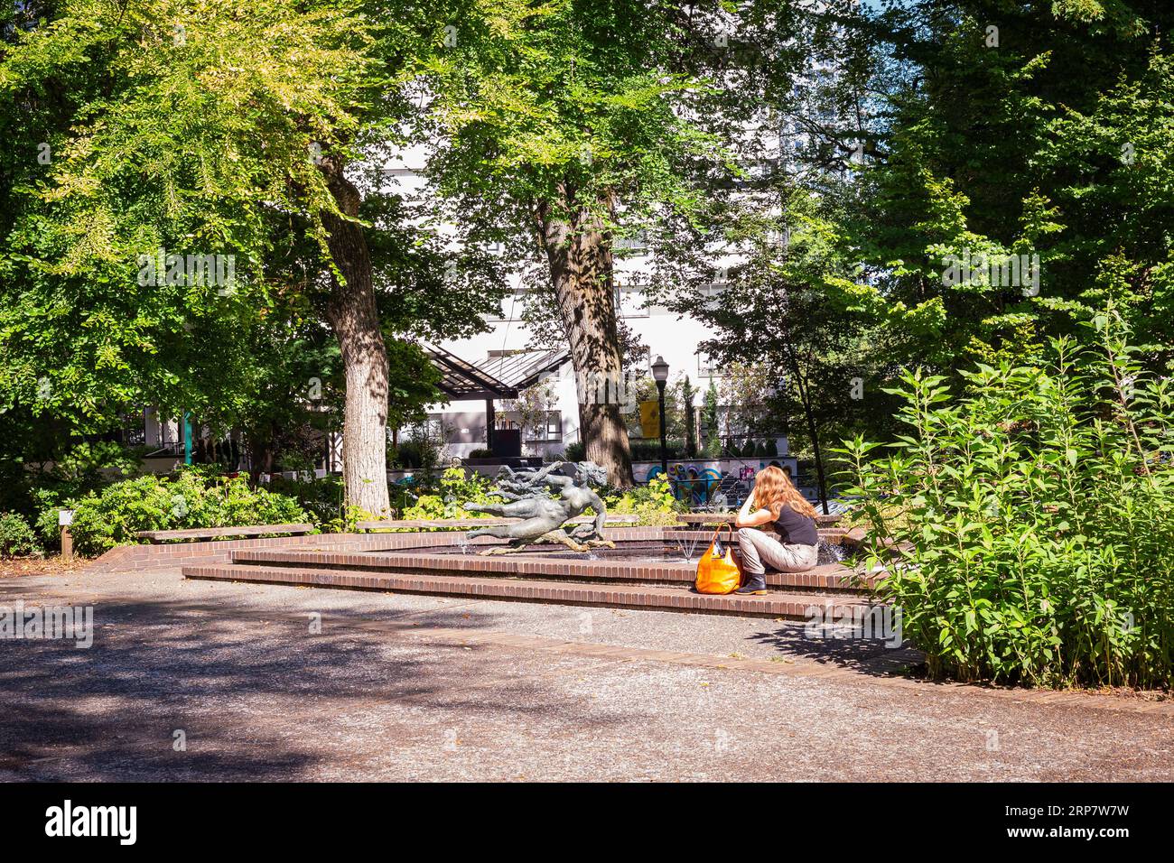 Giovane donna seduta sul bordo di una fontana con una statua nel cortile della Portland State University di Portland, Oregon Foto Stock