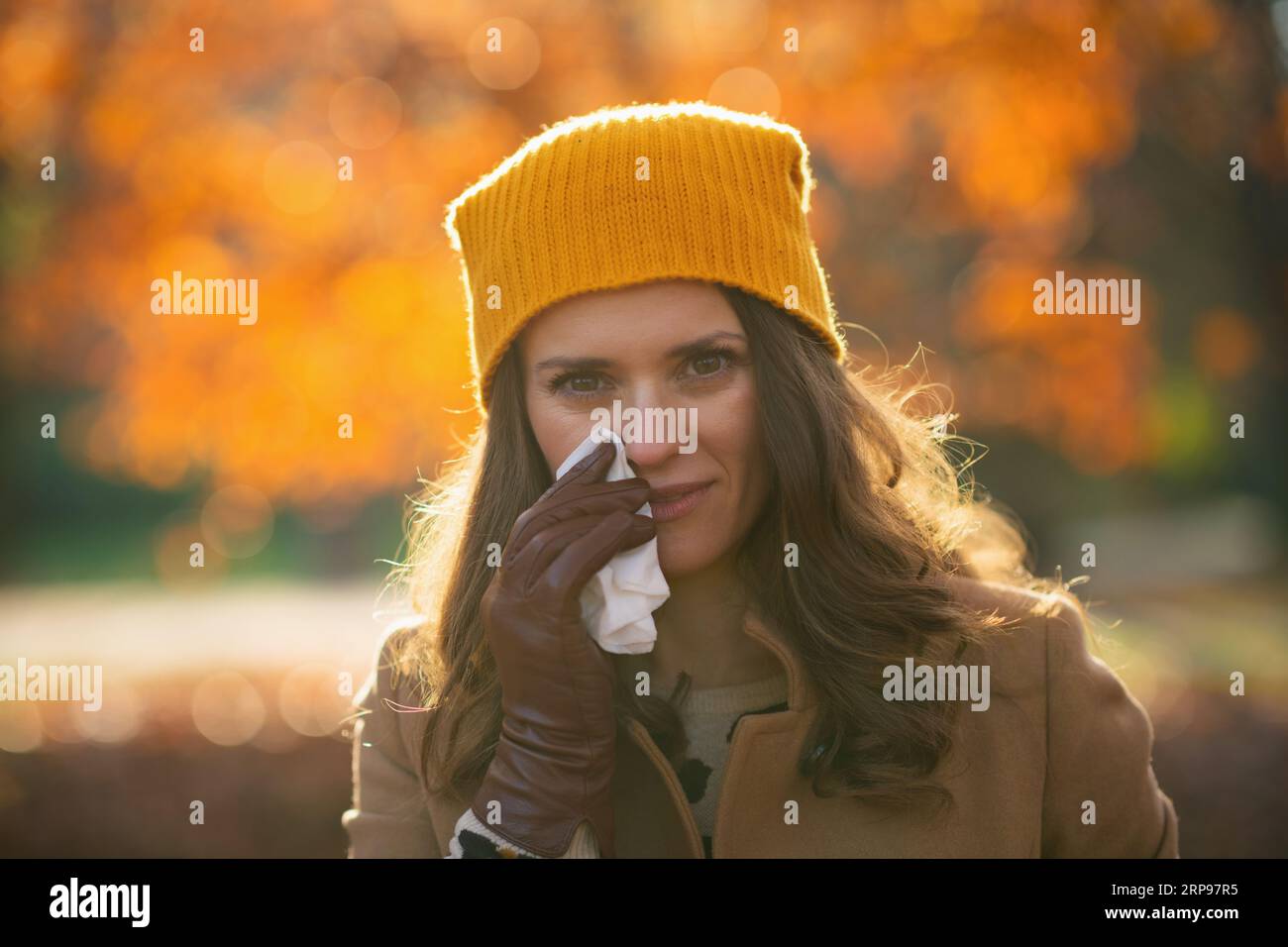 Ciao november. donna alla moda con cappotto marrone e cappello giallo con tovagliolo che soffia naso all'esterno nel parco cittadino in autunno. Foto Stock