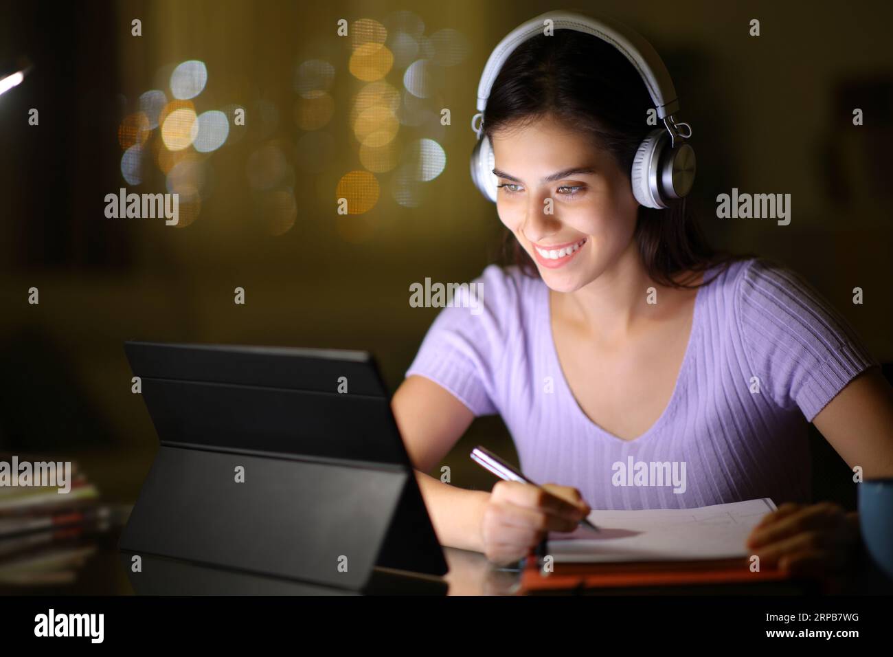 Studente felice che utilizza tablet e cuffie e prende appunti la notte a casa Foto Stock