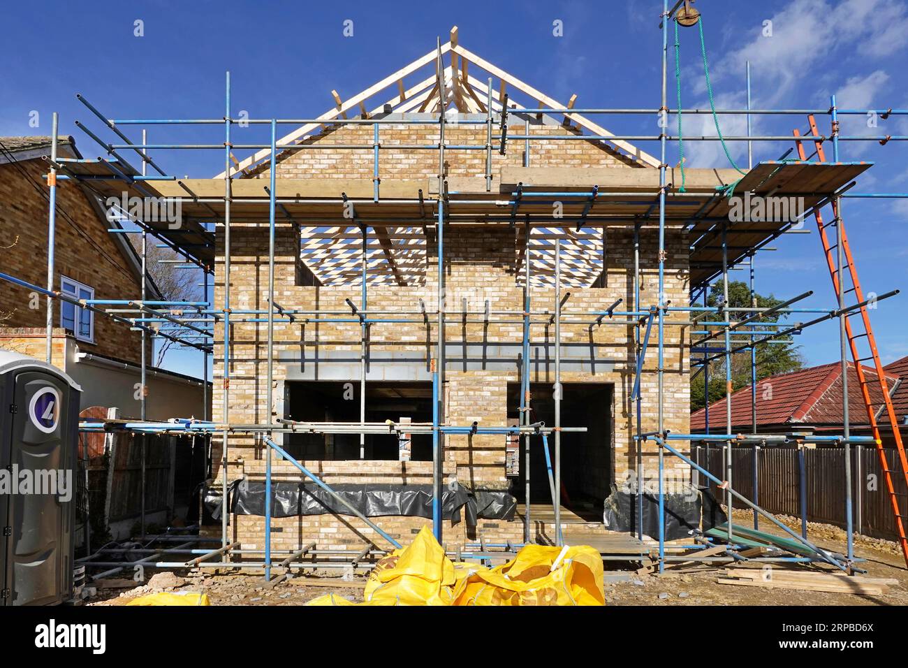 La casa indipendente in mattoni di nuova costruzione con elevazione anteriore sul terreno di riempimento prende forma a parete a timpano e tetto in costruzione avvolto in impalcatura Essex Inghilterra Regno Unito Foto Stock