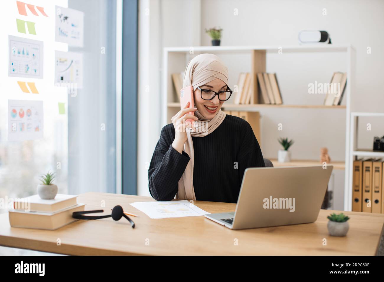 Attraente persona araba che effettua chiamate importanti tramite telefono cellulare mentre scorri le pagine Web sul laptop sul posto di lavoro. Felice imprenditore nella negoziazione hijab Foto Stock