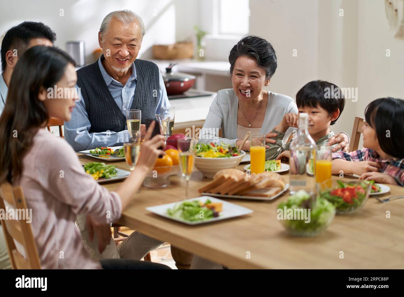 giovane donna asiatica che condivide le foto nel cellulare con una famiglia di tre generazioni mentre pranza insieme Foto Stock