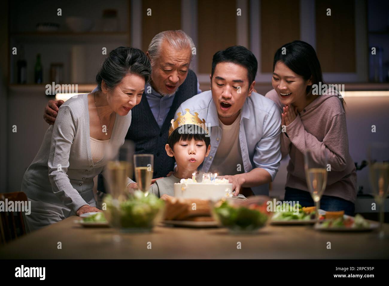 piccolo ragazzo asiatico che soffia candele mentre tre generazioni di famiglia festeggiano il suo compleanno a casa Foto Stock