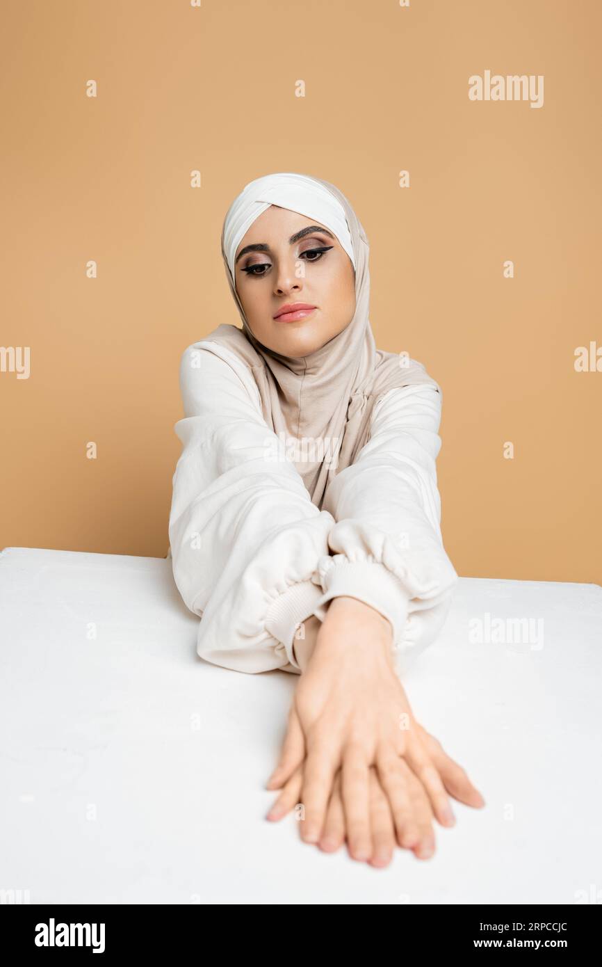 penosa donna mediorientale in hijab e felpa seduto al tavolo bianco su beige, bellezza musulmana Foto Stock