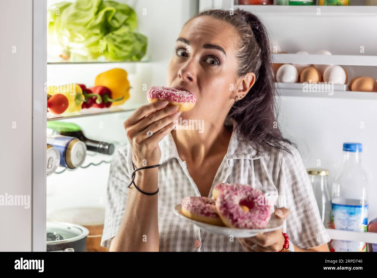 La bruna affamata in pigiama ama ama le ciambelle dolci a tarda notte accanto al frigorifero aperto. Foto Stock