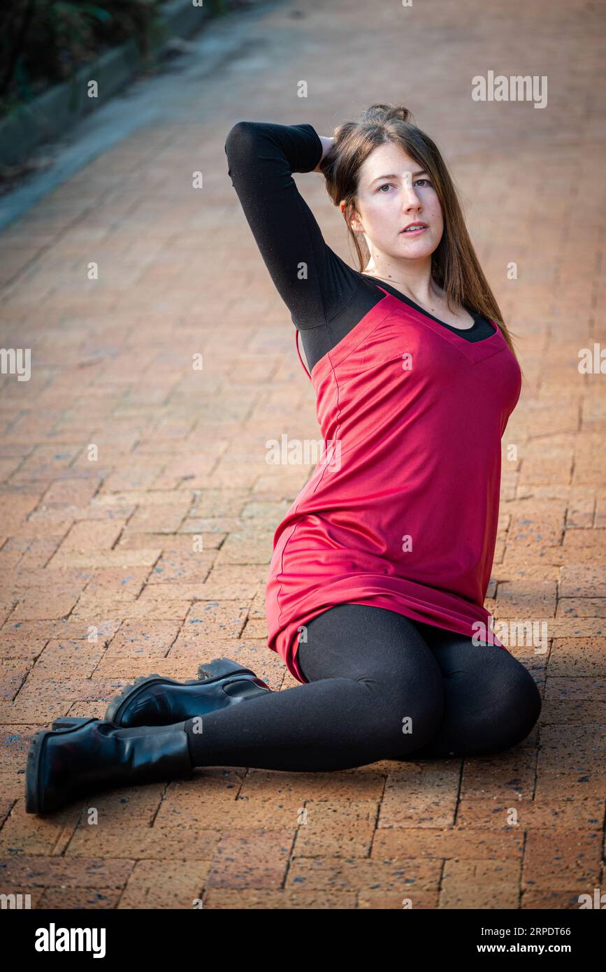 Una bella giovane donna che indossa un vestito rosso e calze nere seduto su un sentiero in mattoni in una posa casual Foto Stock