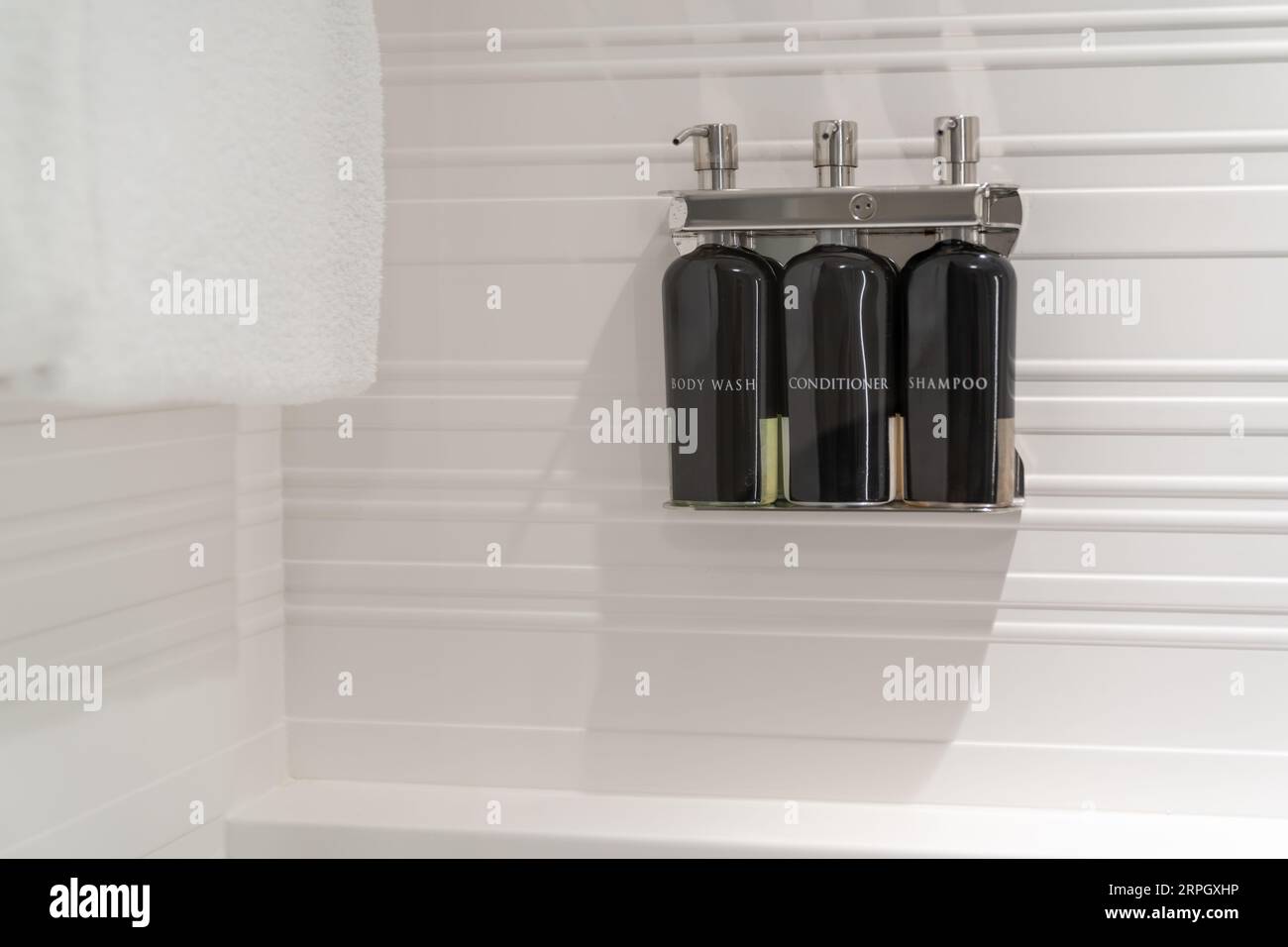 Parete bianca della doccia dell'hotel con bottiglie di detergente per il corpo, sapone liquido, balsamo per capelli e shampoo in un dispenser cromato con un asciugamano (servizi dell'hotel). Foto Stock