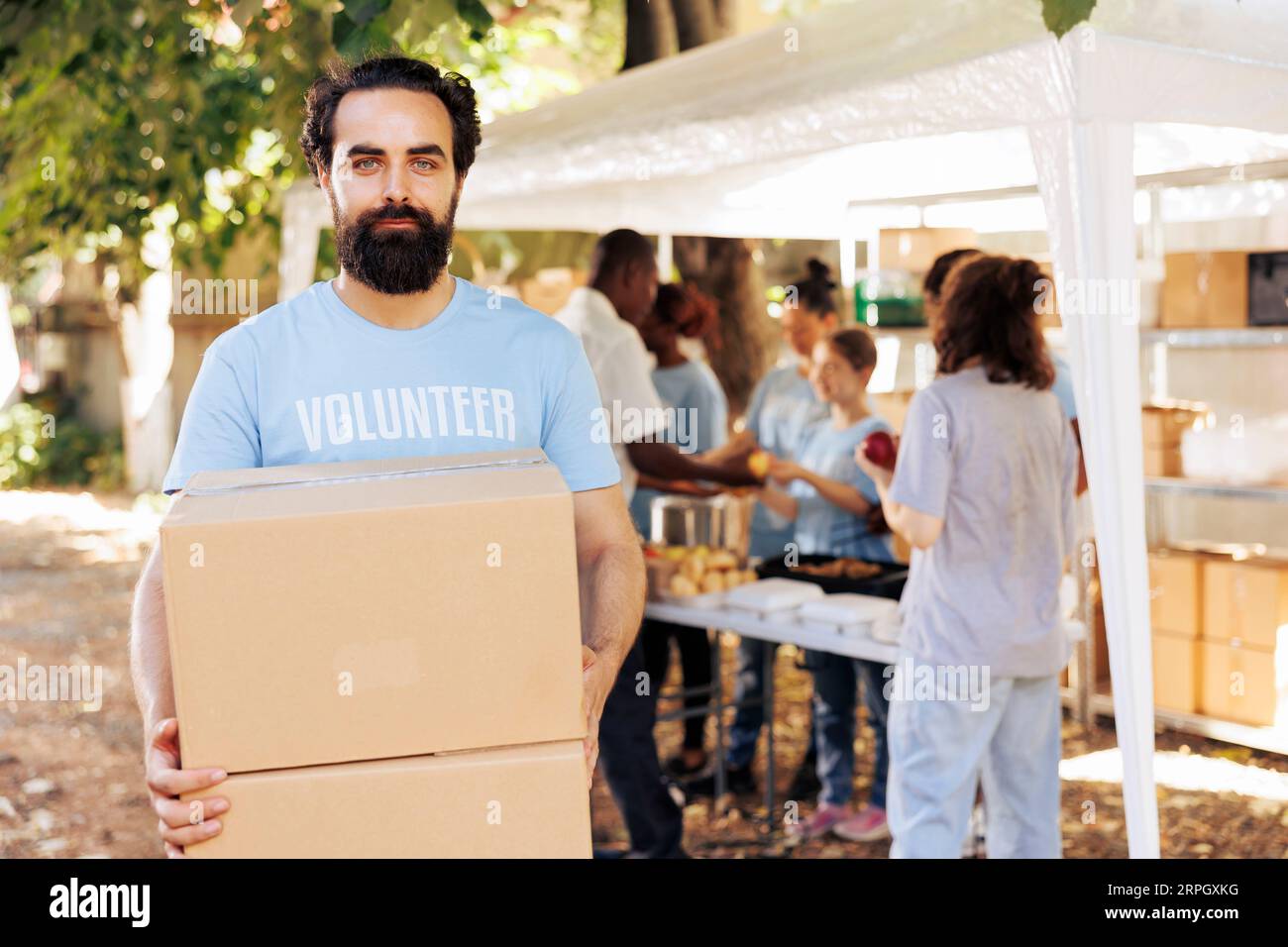 Uomo compassionevole che si offre volontario in un locale food drive, offrendo razioni non deperibili ai bisognosi e ai senzatetto. Lavoratore di beneficenza maschile caucasico che guarda la macchina fotografica mentre trasporta scatole per donazioni Foto Stock