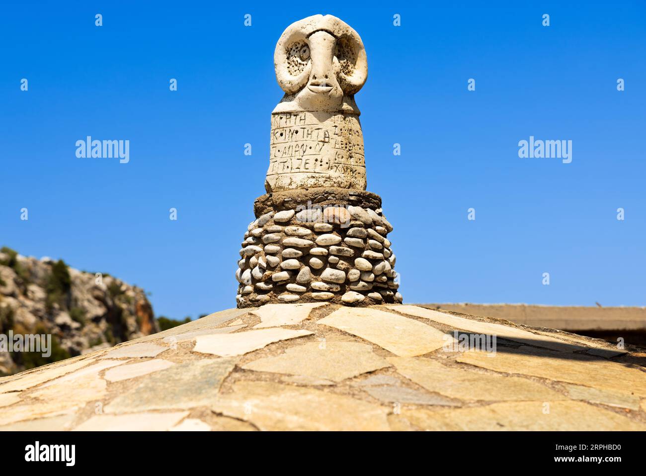Scopri la saggezza senza tempo impressa nello sguardo sereno della statua della testa di un ariete, che adorna con orgoglio il tetto di un santuario cretese. Sotto di esso, l'antico GRE Foto Stock