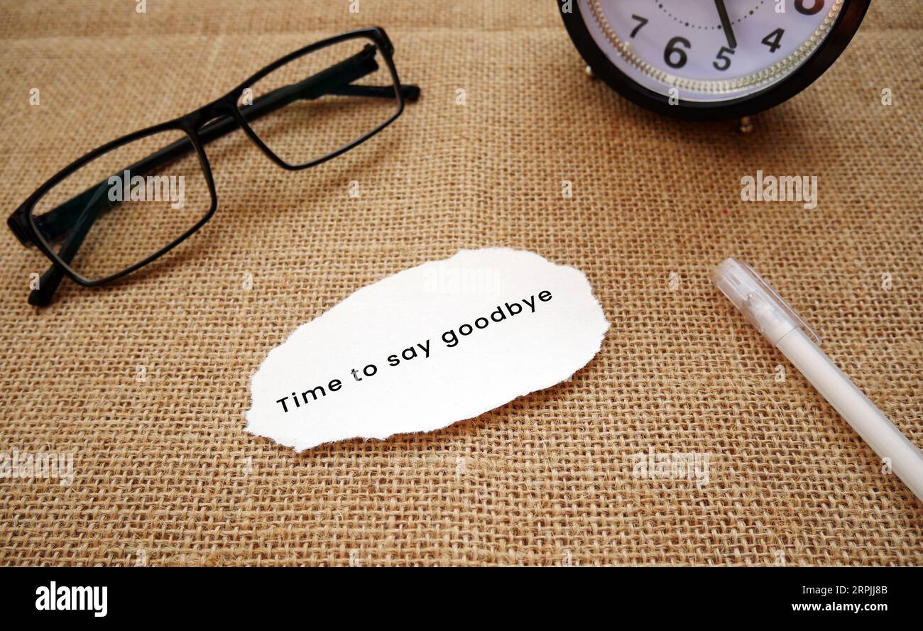 Un orologio sveglia, occhiali, penna e un pezzo di carta color crema strappato che dice il momento di dire addio! su sfondo burlap. Foto Stock