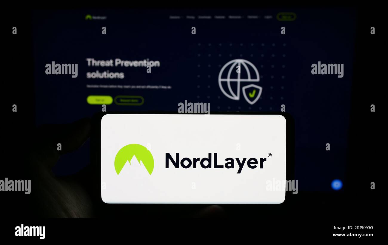 Persona che detiene il cellulare con il logo del servizio di sicurezza per l'accesso alla rete NordLayer sullo schermo di fronte alla pagina Web aziendale. Concentrarsi sul display del telefono. Foto Stock