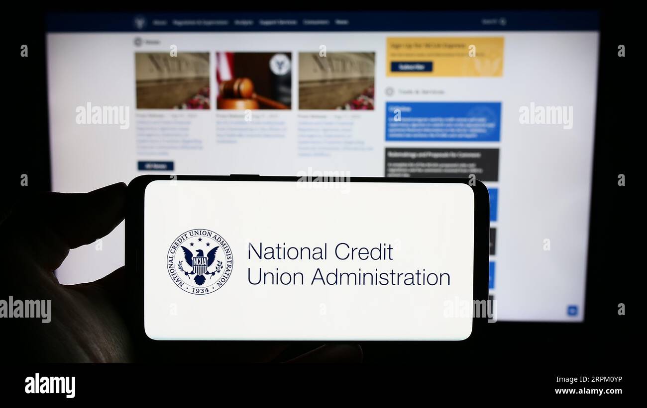Persona in possesso di cellulare con sigillo della National Credit Union Administration (NCUA) sullo schermo davanti alla pagina web. Concentrarsi sul display del telefono. Foto Stock