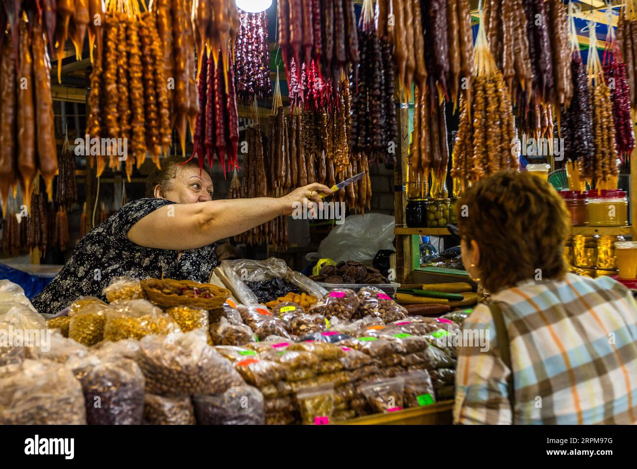 Una merciaia indica con un coltello la churchkhela (georgiano ჩურჭხელა) al suo banco nel mercato coperto dei contadini su Chavchavadze Avenue a Telavi, Georgia. Sorride a un cliente con una camicia a quadri. I filoni sono disposti vicini in arancione, marrone, rosso e viola. Molti terminano con una punta a forma di goccia. Noci e frutta secca in sacchi sono appoggiati sul bancone, con barattoli di conserve a destra. Per preparare questo dolce tradizionale georgiano, vengono infilate noci o nocciole su uno spago. I filoni vengono immersi più volte nel pelamushi, succo d'uva bollito e addensato con farina, e poi essiccati. Chu Foto Stock