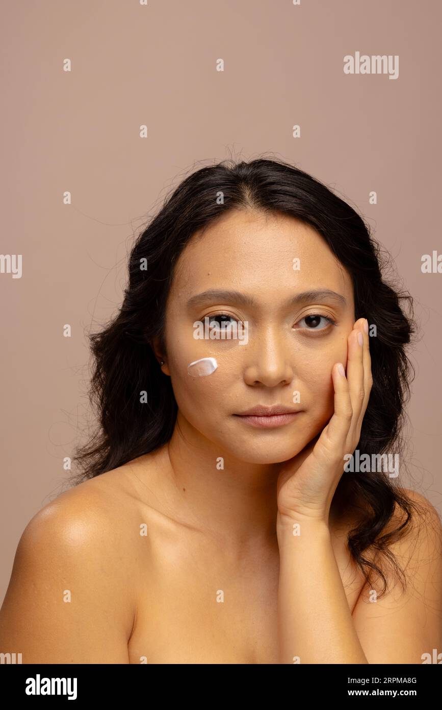 Donna asiatica con capelli scuri con crema per la pelle sulla guancia che tocca il viso con la mano, copia spazio Foto Stock