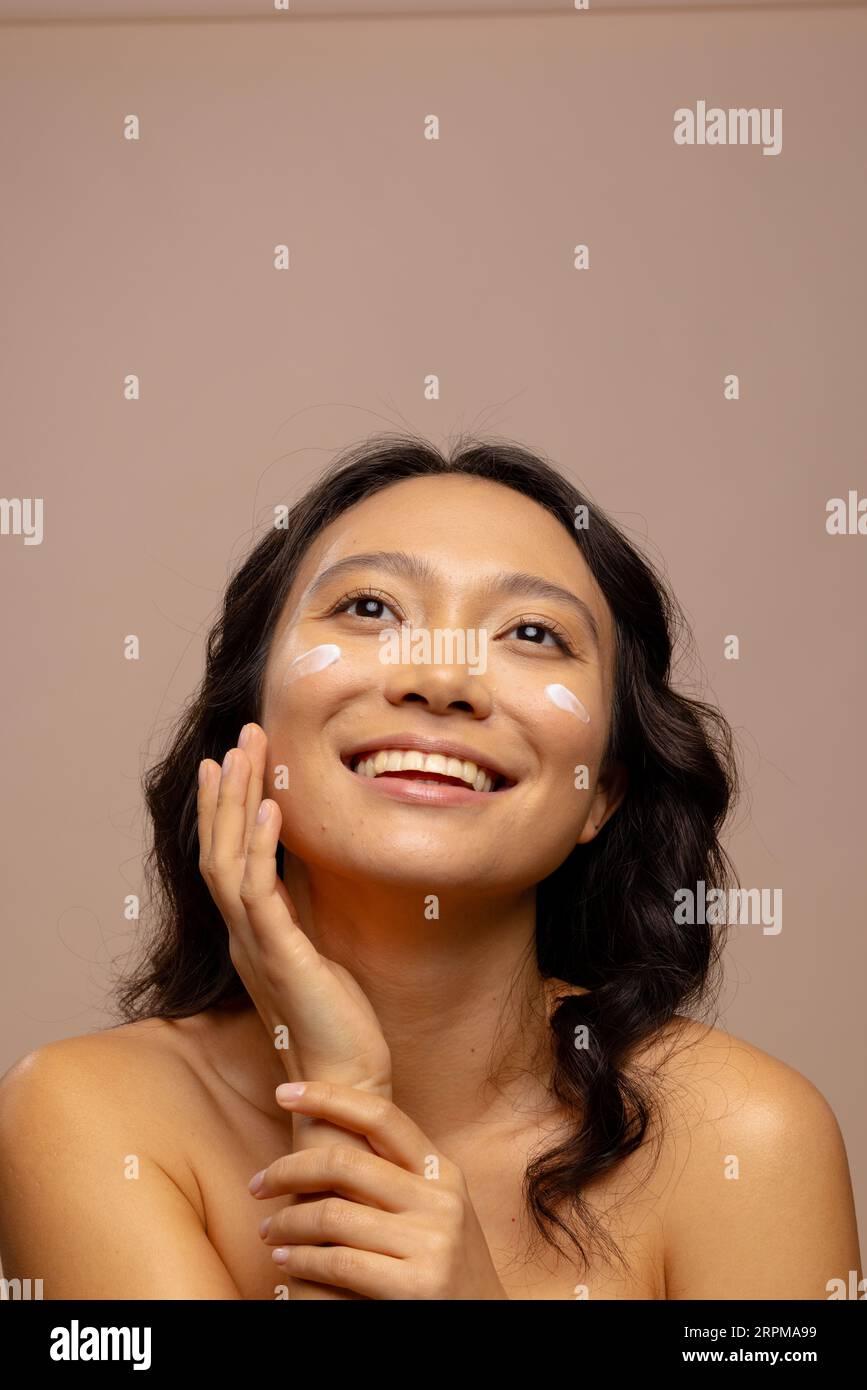 Donna asiatica sorridente con capelli scuri con crema per la pelle sulle guance che tocca il viso, copia spazio Foto Stock
