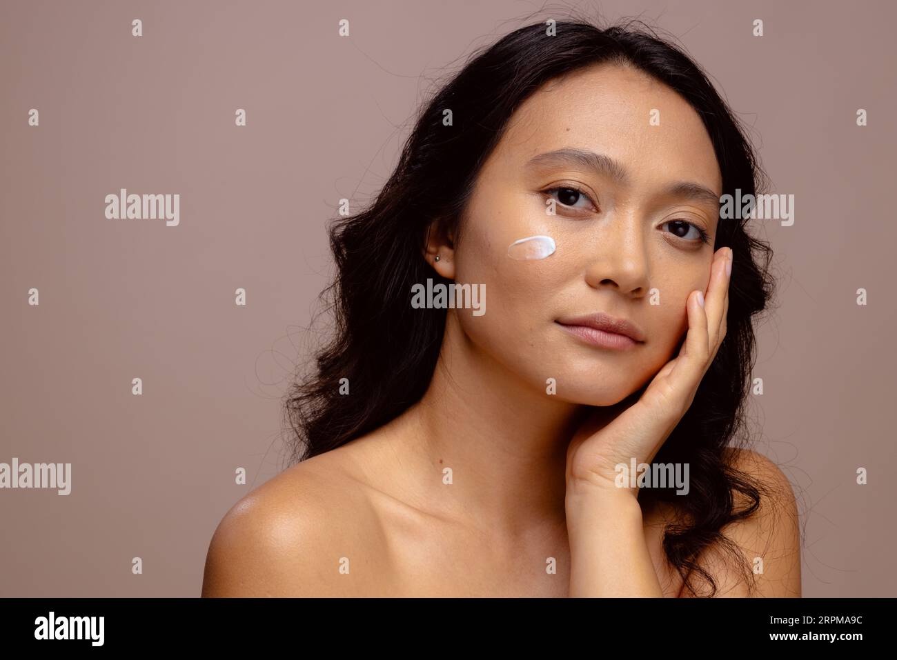 Donna asiatica con capelli scuri con crema per la pelle sulla guancia che tocca il viso con la mano, copia spazio Foto Stock