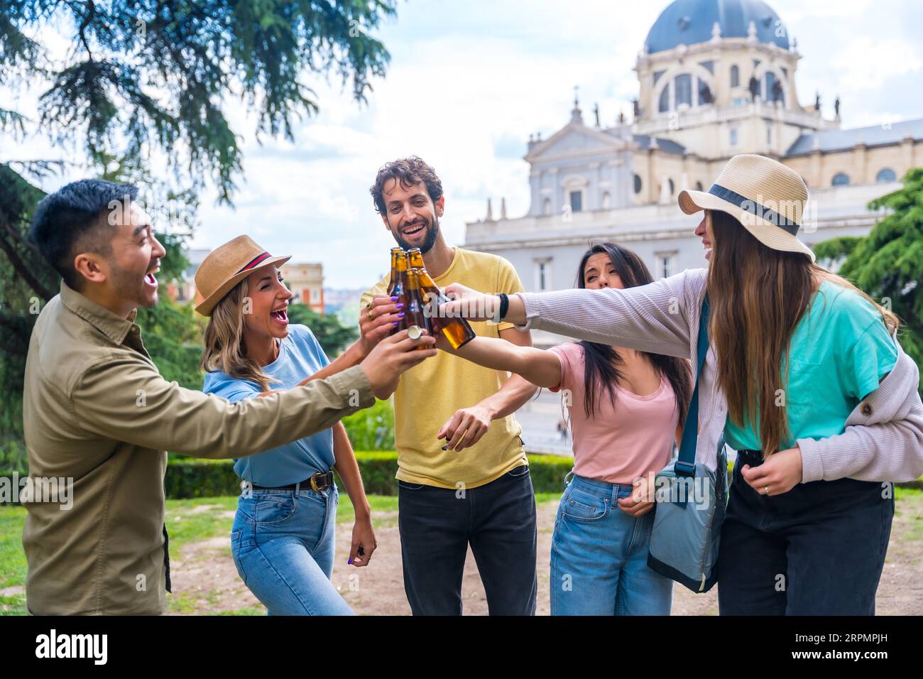Gruppo multietnico di amici che festeggiano in un parco cittadino con birre. Sorridere e tostare con i biberon Foto Stock