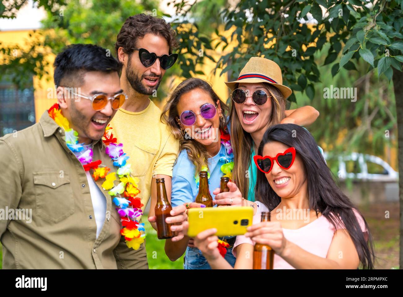 Gruppo multietnico di amici che festeggiano nel parco cittadino scattando un selfie e sorridendo guardando le foto al telefono Foto Stock
