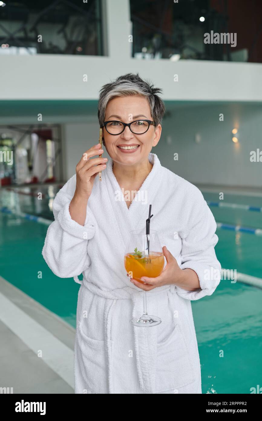 concetto spa benessere, donna felice e matura in bicchieri che tiene un cocktail e parla sullo smartphone Foto Stock