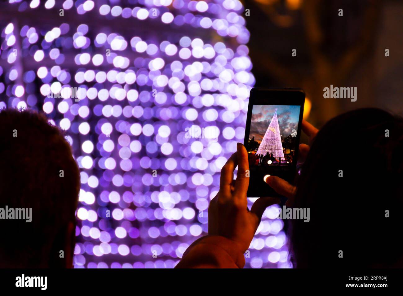 Silhouette retroilluminata di una persona non identificabile che scatta una foto con il suo cellulare della decorazione natalizia per strada Foto Stock