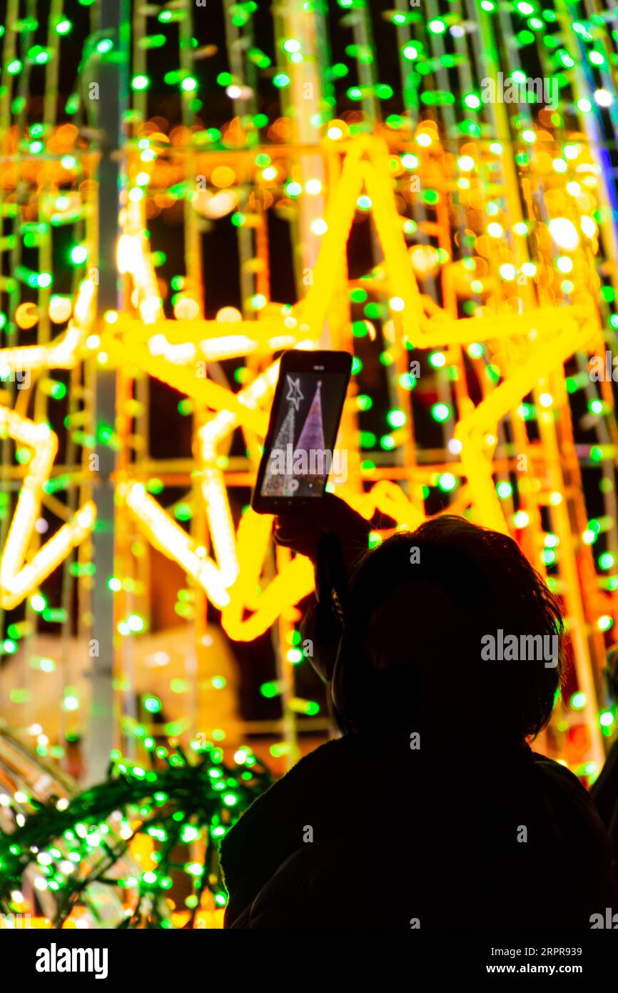Silhouette retroilluminata di una persona non identificabile che scatta una foto con il suo cellulare della decorazione natalizia per strada Foto Stock