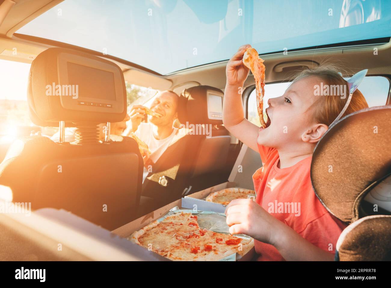 Bambina con ritratto a bocca aperta mangiare pizza italiana seduta in macchina moderna con madre e padre. Momenti felici in famiglia, infanzia, fast food mangiare Foto Stock