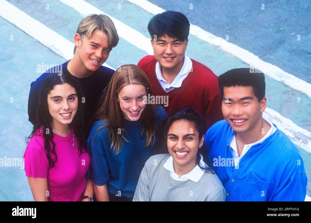 Studenti scolastici 15-17 anni gruppo etnico multiculturale composto da 6 giovani felici e attraenti studenti della scuola superiore del liceo multiculturale in magliette colorate si sfidano e si mettono in posa all'aperto in un ambiente soleggiato del campus universitario. Studenti adolescenti ispanici, coreani, asiatici, orientali Foto Stock