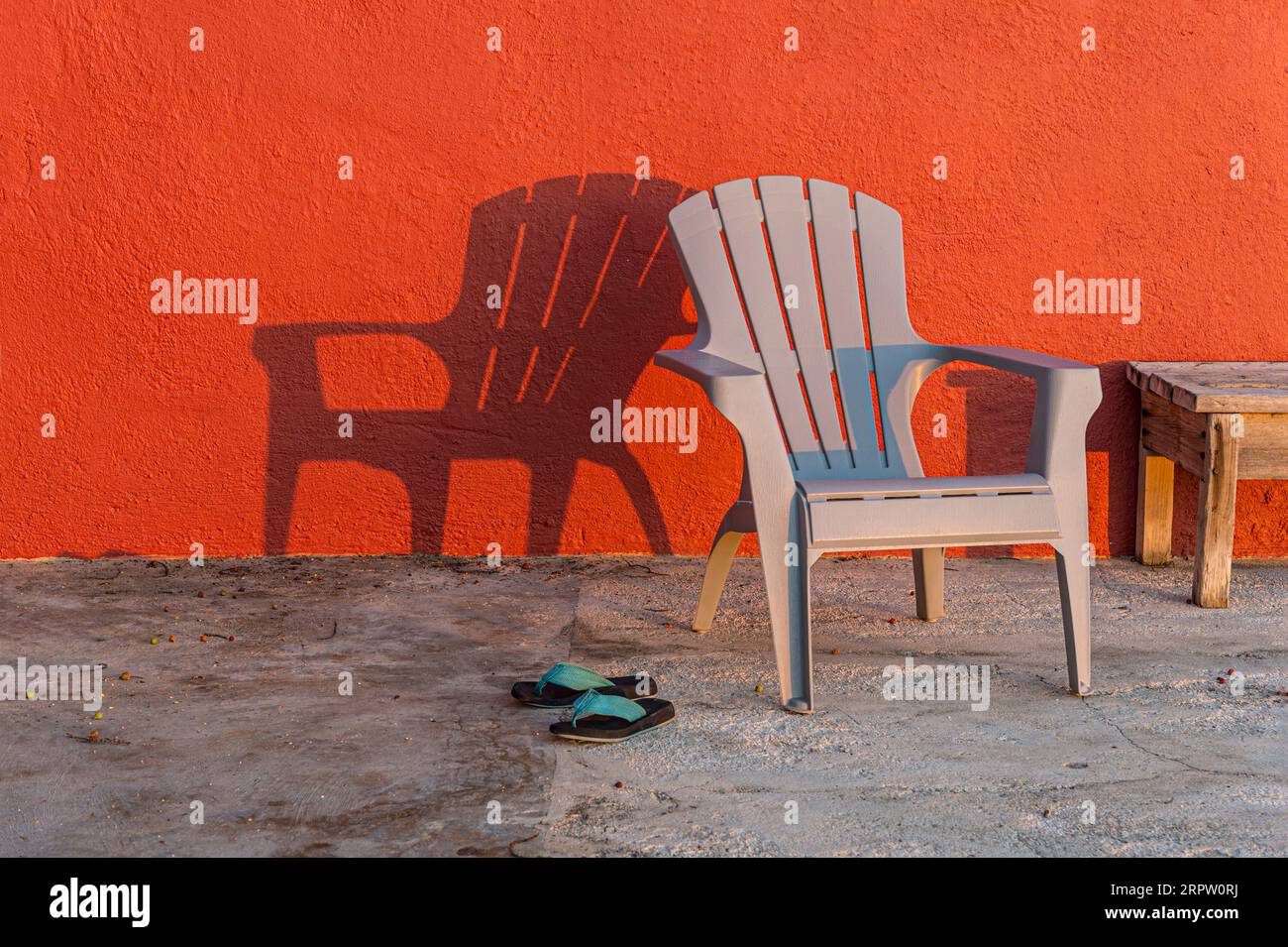 Sedia solitaria al sole nel tardo pomeriggio con parete rossa e scarpe Foto Stock