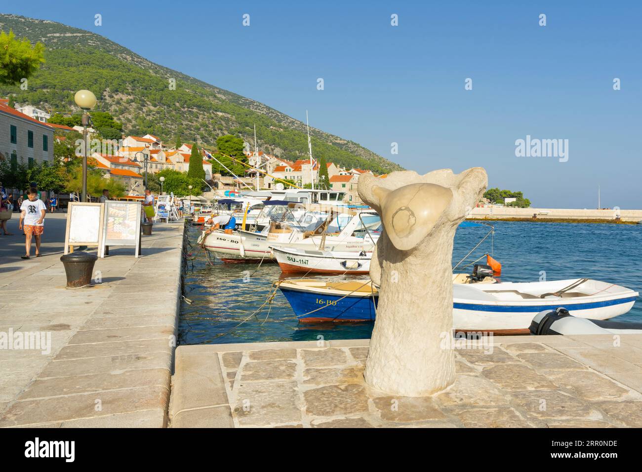 Croazia Island Brac Bol porto città sul lungomare, pietra astratta scultura, statue, ormeggi d'arte barche a motore, bar ristorante per turisti Foto Stock
