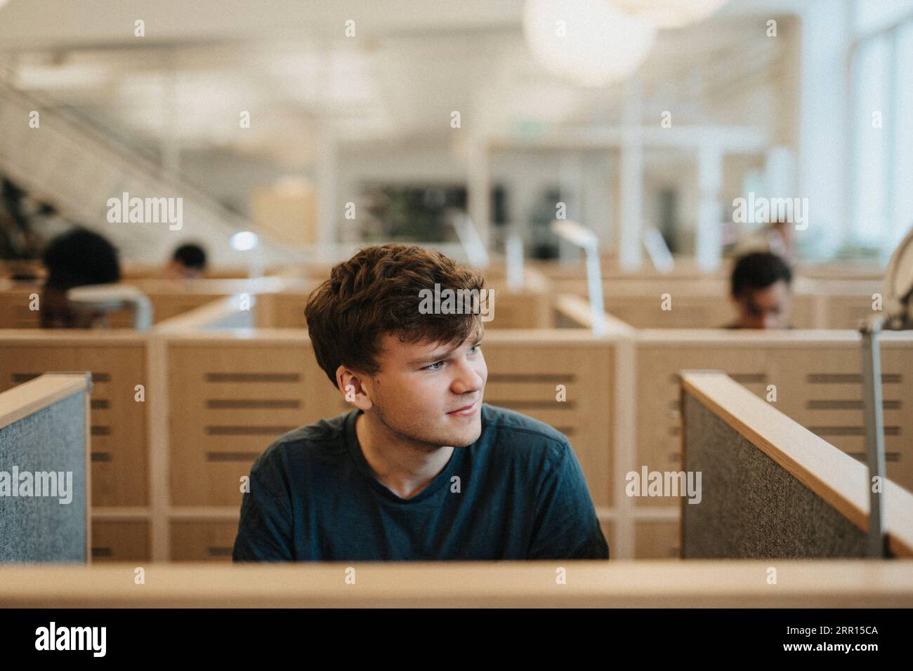 Un giovane sorridente che guarda lontano mentre è seduto in biblioteca all'università Foto Stock