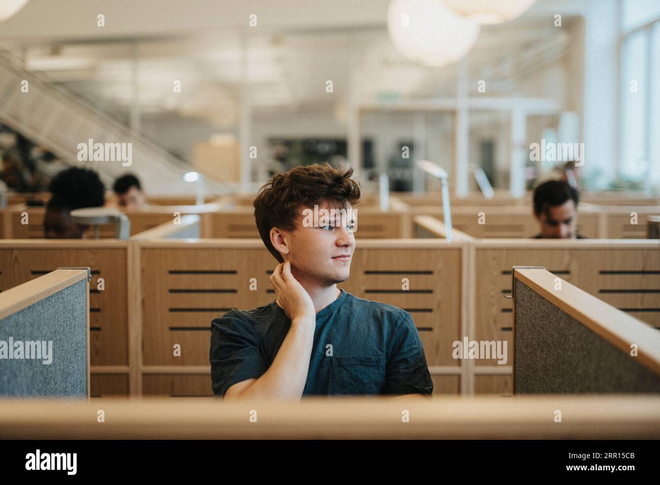 Uno studente maschio sorridente che guarda lontano mentre è seduto in biblioteca all'università Foto Stock