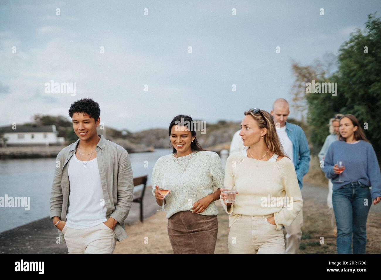 Giovani amici maschi e femmine che bevono mentre camminano insieme vicino al lago Foto Stock