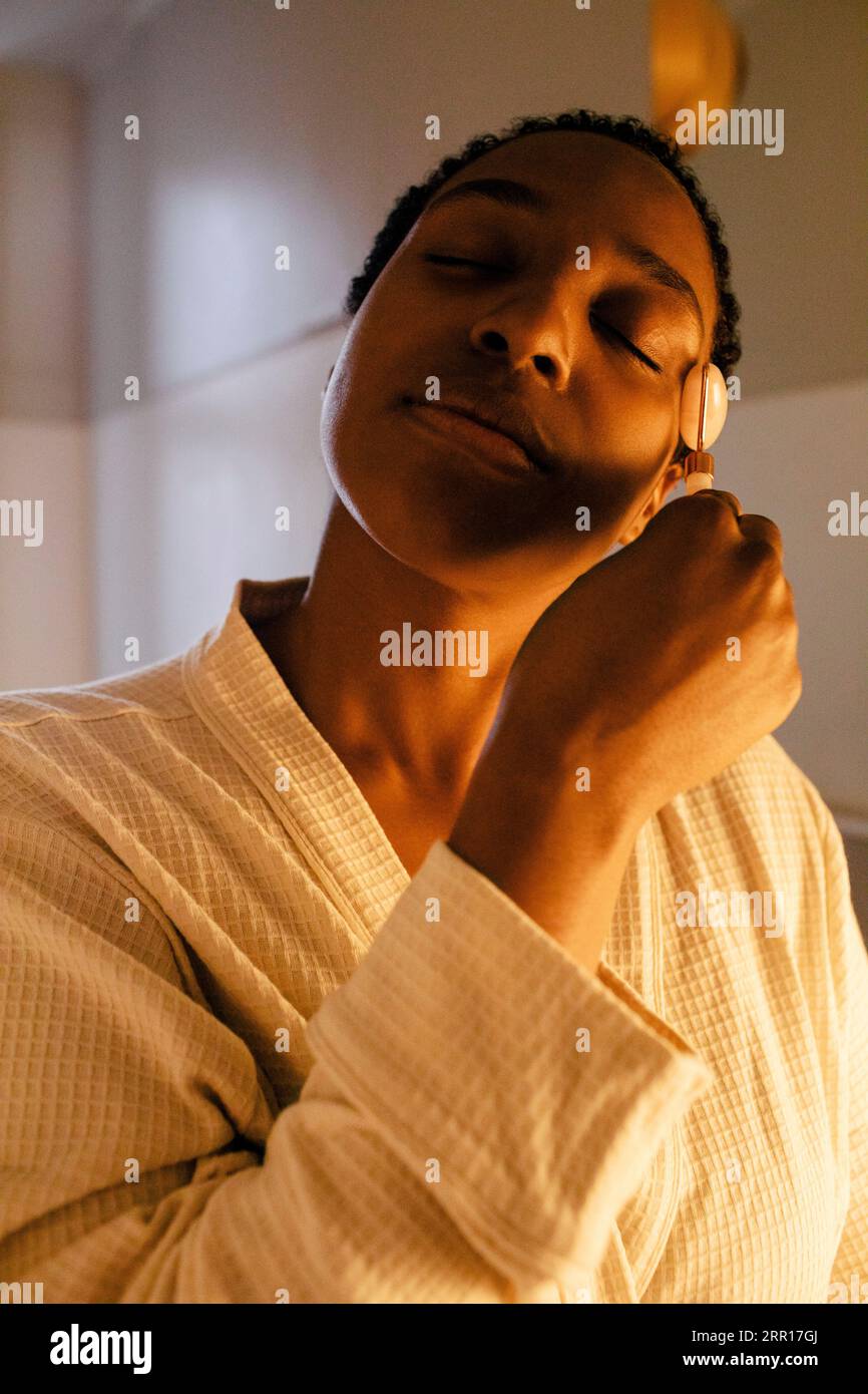 Donna con gli occhi chiusi che usa il rullo di giada sul viso in bagno Foto Stock