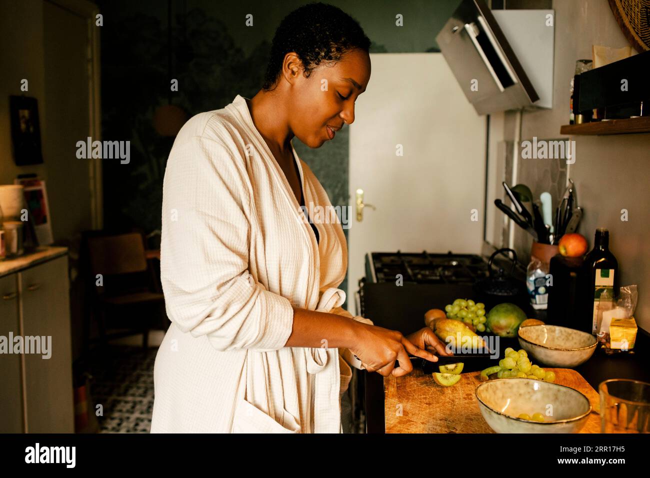Vista laterale di una giovane donna che taglia kiwi al bancone della cucina Foto Stock