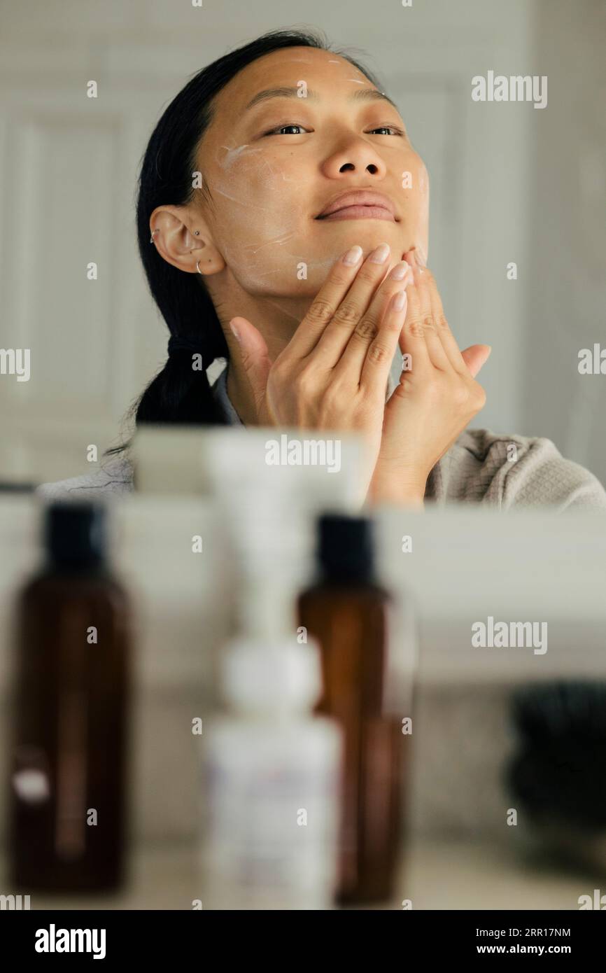 Riflesso della giovane donna che applica idratante sul viso a casa Foto Stock