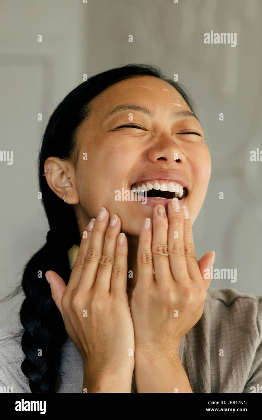 Giovane donna felice che applica idratante sul viso a casa Foto Stock