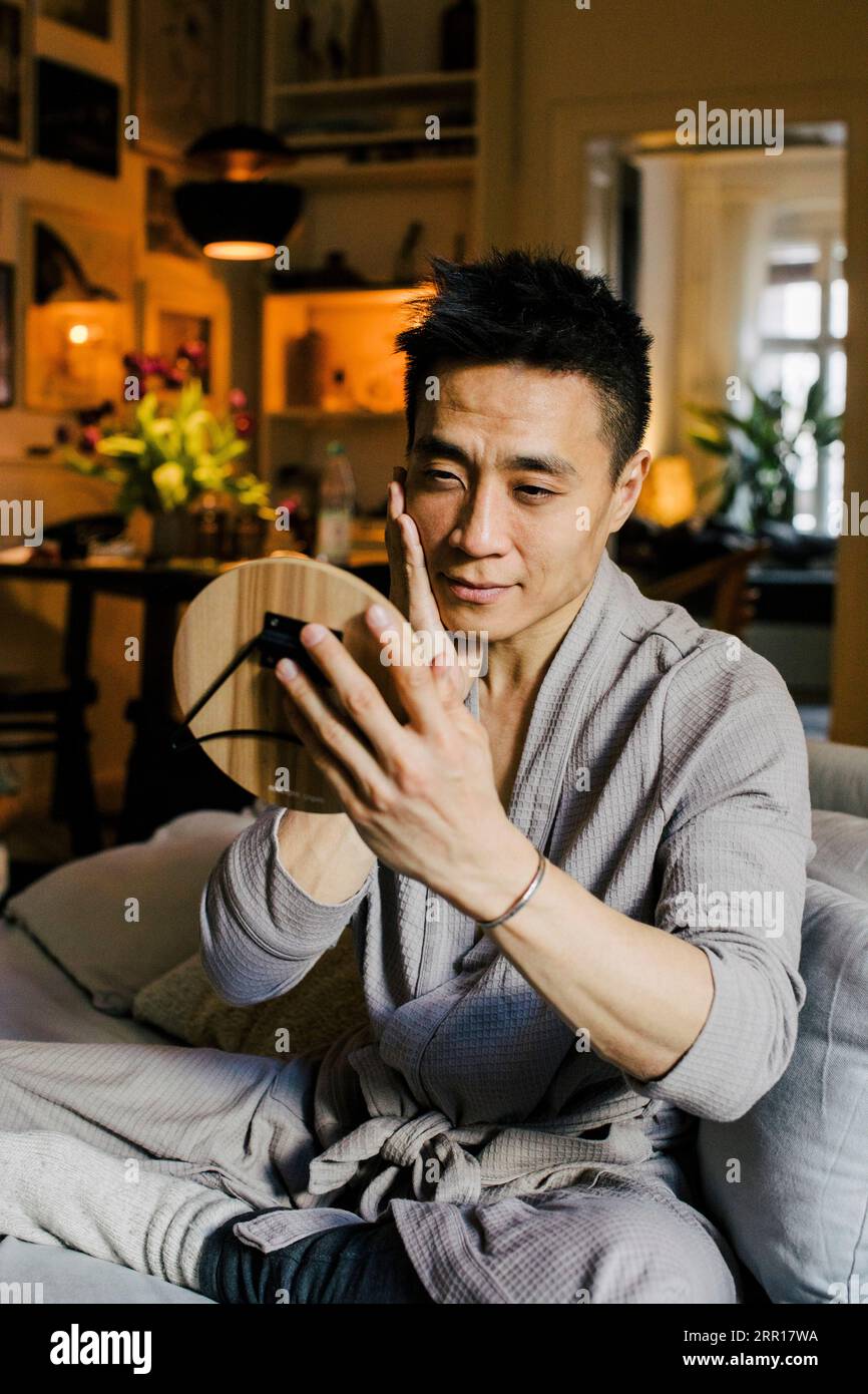 Uomo che guarda allo specchio mentre è seduto sul divano a casa Foto Stock