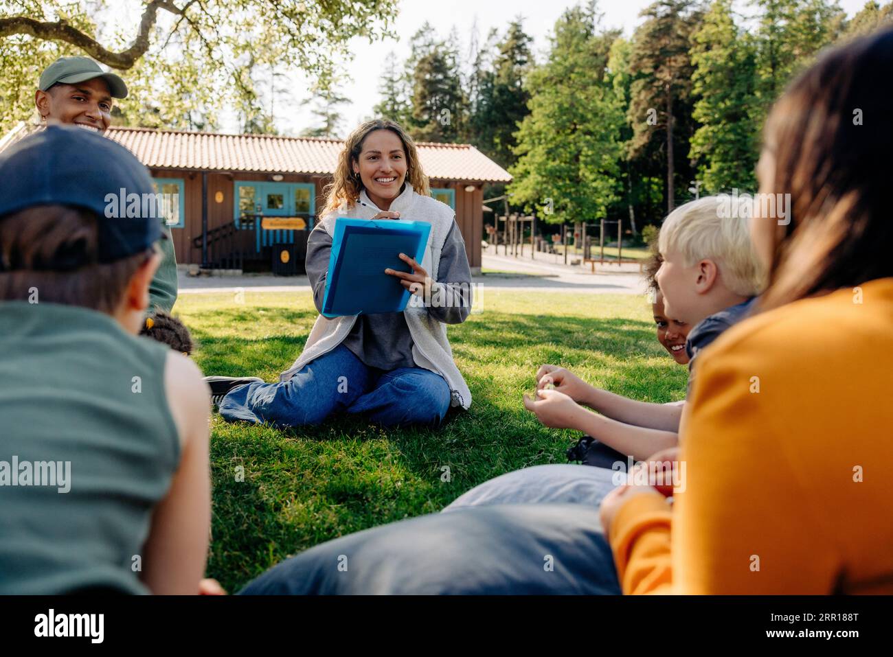 Consiglieri sorridenti che fanno attività con bambini seduti sull'erba al campo estivo Foto Stock